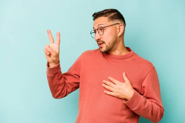 Genç İspanyol bir adam mavi arka planda izole edilmiş yemin ediyor, elini göğsüne koyuyor..
