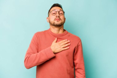 Genç İspanyol bir adam mavi arka planda izole edilmiş yemin ediyor, elini göğsüne koyuyor..