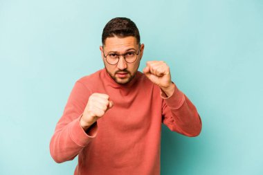 Latin kökenli genç bir adam mavi arka planda izole edilmiş. Yumruk atıyor, sinirleniyor, kavga ediyor..