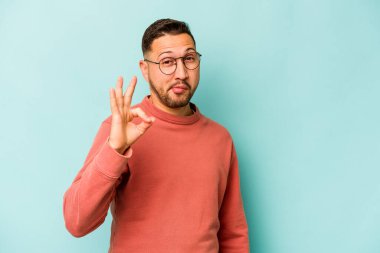 Mavi arka planda izole edilmiş genç İspanyol bir adam bir göz kırptı ve elleriyle iyi bir jest yaptı..