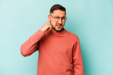 Genç İspanyol bir adam mavi arka planda izole edilmiş parmaklarıyla kulaklarını kapatıyor, gürültülü bir ortam yüzünden stresli ve çaresiz..