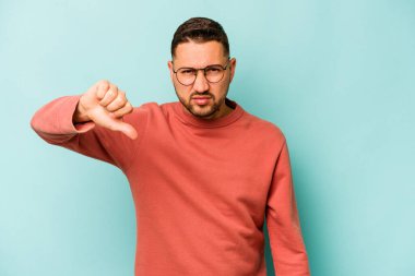 Mavi arka planda izole edilmiş genç İspanyol adam baş parmağını aşağı gösteriyor ve hoşlanmadığını ifade ediyor..