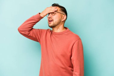 Mavi arka planda izole edilmiş genç İspanyol adam elleriyle gözlerini kapatıyor, geniş bir gülümsemeyle bir sürpriz bekliyor..