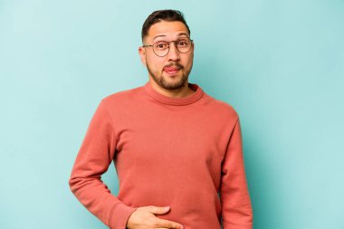 Mavi arka planda izole edilmiş genç İspanyol adam karnına dokunuyor, nazikçe gülümsüyor, yemek yiyor ve tatmin oluyor..