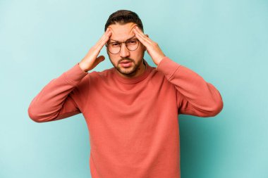 Genç İspanyol adam mavi arka planda izole edilmiş tapınaklara dokunuyor ve baş ağrısı çekiyor..