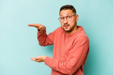 Mavi arka planda izole edilmiş genç İspanyol adam iki eliyle bir şey tutuyor, ürün sunumu..