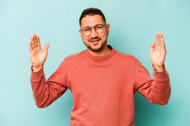 Mavi arka planda izole edilmiş genç İspanyol adam işaret parmaklarıyla küçük bir şey tutuyor, gülümsüyor ve kendinden emin..