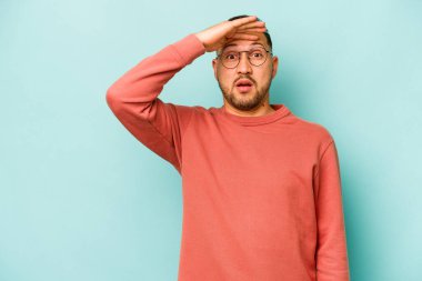 Genç İspanyol adam mavi arka planda izole edilmiş, uzaklara bakıyor ve elini alnına koyuyor..