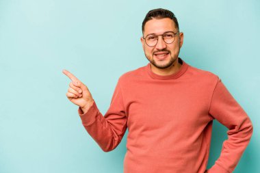 Mavi arka planda izole edilmiş genç İspanyol adam işaret parmağıyla işaret ederek gülümsüyor..