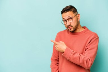 Mavi arka planda izole edilmiş genç İspanyol adam gülümsüyor ve boş alanda bir şeyler gösteriyor..