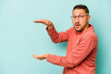 Mavi arka planda izole edilmiş genç İspanyol adam elleri arasında bir kopya alanı tutarken şok oldu ve hayrete düştü..