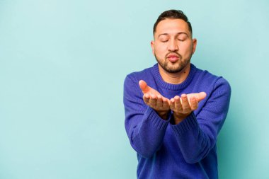 Genç İspanyol adam mavi arka planda izole edilmiş dudaklar kıvrılıyor ve hava öpücüğü göndermek için avuç içleri tutuyor..