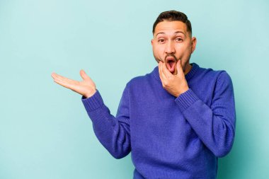 Mavi arka planda izole edilmiş genç İspanyol bir adam avucunun içinde fotokopi alanı tutuyor, el üstünde tutuyor. Şaşırmış ve mutlu..