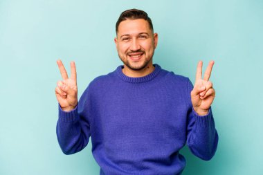 Mavi arka planda izole edilmiş genç İspanyol adam zafer işareti gösteriyor ve gülümsüyor..