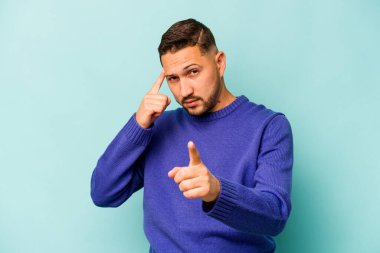 Genç İspanyol bir adam mavi arka planda izole edilmiş, parmağıyla tapınağı işaret ediyor, düşünüyor ve bir göreve odaklanıyor..