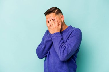 Mavi arka planda izole edilmiş genç İspanyol adam kameraya parmaklarıyla göz kırpıyor, yüzünü kapatmaktan utanıyor..