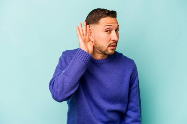 Genç İspanyol bir adam mavi arka planda izole edilmiş bir şekilde bir dedikoduyu dinlemeye çalışıyor..