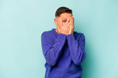 Genç İspanyol adam mavi arka planda izole edilmiş. Korkmuş ve gergin parmaklarıyla göz kırp..