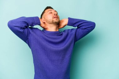 Genç İspanyol adam mavi arka planda izole edilmiş. Kendine güveniyor, elleri başının arkasında..