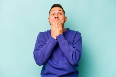 Mavi arka planda izole edilmiş genç İspanyol adam elleriyle ağzını kapatırken şok oldu..