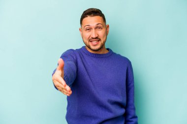 Mavi arka planda izole edilmiş genç İspanyol adam selamlama jesti olarak kameraya uzanıyor..