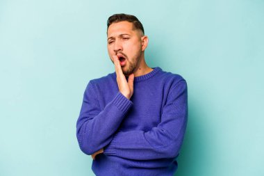 Mavi arka planda izole edilmiş genç İspanyol adam esniyor. Ağzı eliyle kapatan yorgun bir hareket sergiliyor..