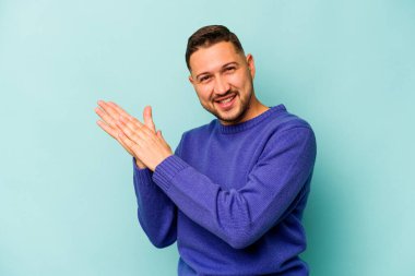 Genç İspanyol adam mavi arka planda izole edilmiş enerjik ve rahat hissediyor, ellerini ovuşturuyor kendinden emin..