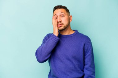 Genç İspanyol adam mavi arka planda izole edilmiş. Çok yorgun ve uykulu. Elini başının üstünde tutuyor..