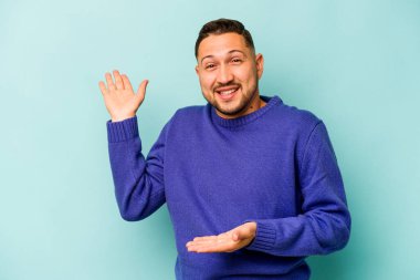 Genç İspanyol bir adam mavi arka planda izole edilmiş. Kafası karışmış ve omuz silkiyor. Kopya alanını tutmak için..