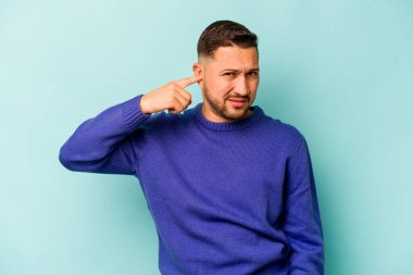 Mavi arka planda izole edilmiş genç İspanyol adam kulaklarını elleriyle kapatıyor..
