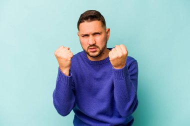 Mavi arka planda izole edilmiş genç İspanyol adam kameraya yumruk gösteriyor, agresif yüz ifadesi..