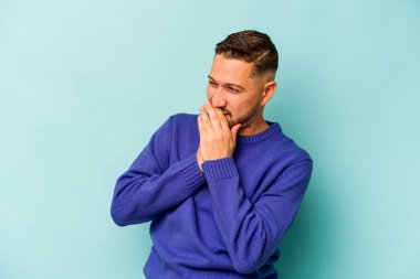 Genç İspanyol bir adam mavi arka planda izole edilmiş bir şekilde bir şeye gülüyor, elleriyle ağzını kapatıyor..