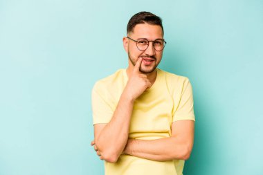 Genç İspanyol adam mavi arka planda izole edilmiş mutlu ve kendinden emin gülümsüyor, elleriyle çenesine dokunuyor..