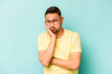 Mavi arka planda izole edilmiş genç İspanyol adam yanakları patlatıyor, yorgun bir ifadesi var. Yüz ifadesi kavramı.