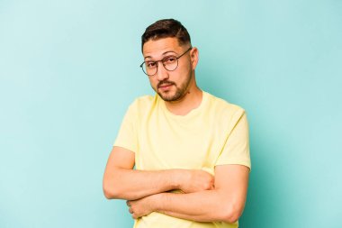 Latin asıllı genç adam mavi arka planda izole edilmiş. Mutsuz bir şekilde kameraya bakıyor. İğneleyici bir ifadeyle..