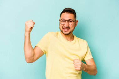 Genç İspanyol adam mavi arka planda izole edilmiş kaygısız ve heyecanlı tezahüratlar yapıyor. Zafer konsepti.