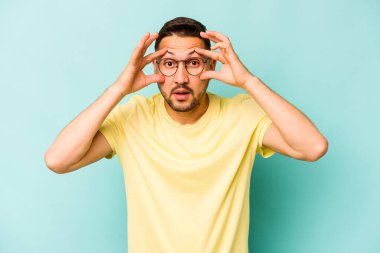 Genç İspanyol bir adam mavi arka planda izole edilmiş. Başarı fırsatı bulmak için gözlerini açık tutuyor..