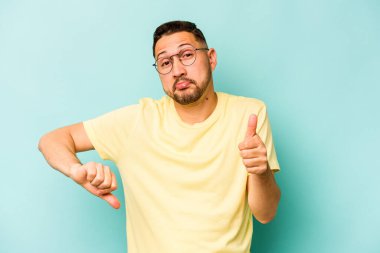 Latin kökenli genç adam mavi arka planda izole edilmiş, baş parmağını kaldırıyor, zor bir seçim konsepti.
