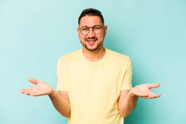 Mavi arka planda izole edilmiş genç İspanyol adam kollarıyla ölçeği yapıyor, mutlu ve kendinden emin hissediyor..