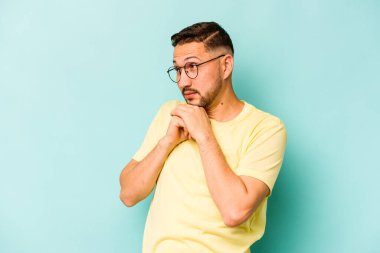 Latin kökenli genç adam mavi arka planda izole edilmiş şans için dua ediyor, şaşırmış ve ağzı açık bir şekilde önüne bakıyor..