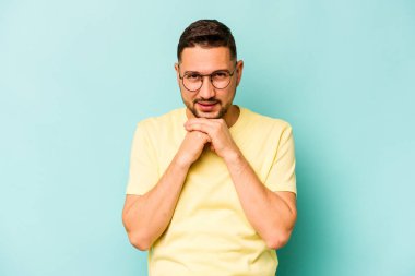 Mavi arka planda izole edilmiş genç İspanyol adam ellerini çenesinin altında tutuyor ve mutlu bir şekilde yan tarafa bakıyor..