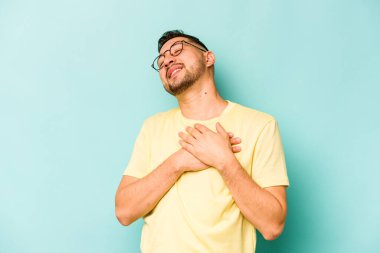 Mavi arka planda izole olmuş genç İspanyol adam gülüyor, ellerini yüreğine koyuyor, mutluluk kavramını koruyor..