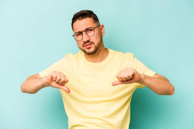 Mavi arka planda izole edilmiş genç İspanyol adam baş parmağını aşağı gösteriyor, hayal kırıklığı konsepti.
