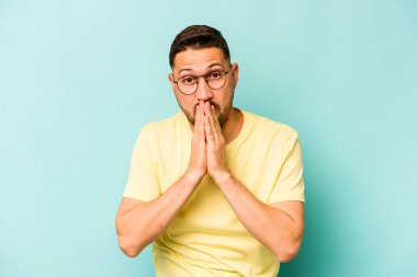 Genç İspanyol bir adam mavi arka planda izole edilmiş, ağzı kapalı ve elleri endişeli görünüyor..
