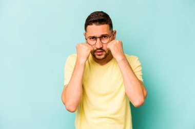 Latin kökenli genç bir adam mavi arka planda izole edilmiş. Yumruk atıyor, sinirleniyor, kavga ediyor..