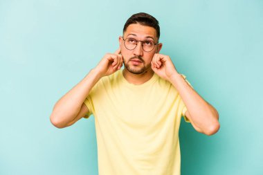 Genç İspanyol bir adam mavi arka planda izole edilmiş parmaklarıyla kulaklarını kapatıyor, gürültülü bir ortam yüzünden stresli ve çaresiz..