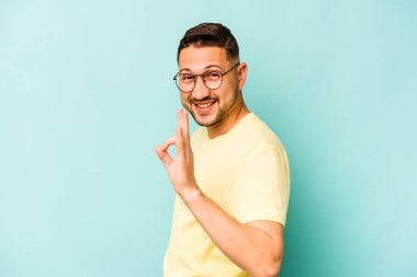 Mavi arka planda izole edilmiş genç İspanyol bir adam bir göz kırptı ve elleriyle iyi bir jest yaptı..