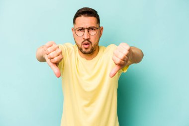 Mavi arka planda izole edilmiş genç İspanyol adam baş parmağını aşağı gösteriyor ve hoşlanmadığını ifade ediyor..