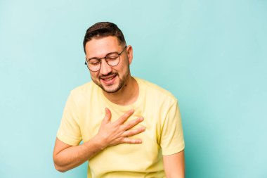 Mavi arka planda izole edilmiş genç İspanyol adam yüksek sesle gülüyor ve elini göğsüne koyuyor..