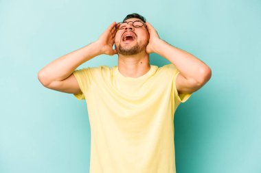 Mavi arka planda izole edilmiş genç İspanyol adam ellerini başının üstünde tutarak neşeyle gülüyor. Mutluluk kavramı.